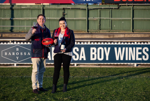 Two people standing on a grassy field in front of a large blue and white B Barossa & Barossa Boy Wines sign, holding wine bottles and a Sherrin football.